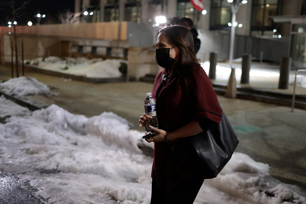 A dark-haired woman carrying a water bottle and with a bag slung over her should, leaves a building and walks on snow-covered pavement.