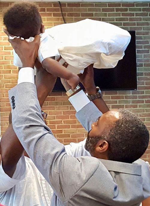 Two men hold a baby in white above their heads