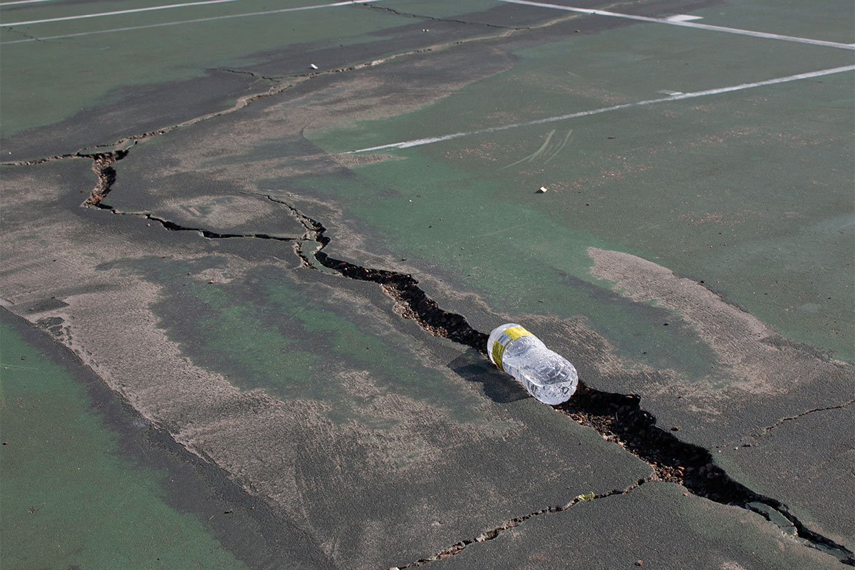 A wide crack is seen on the ground of a sport area, plastic bottle debris in the crack