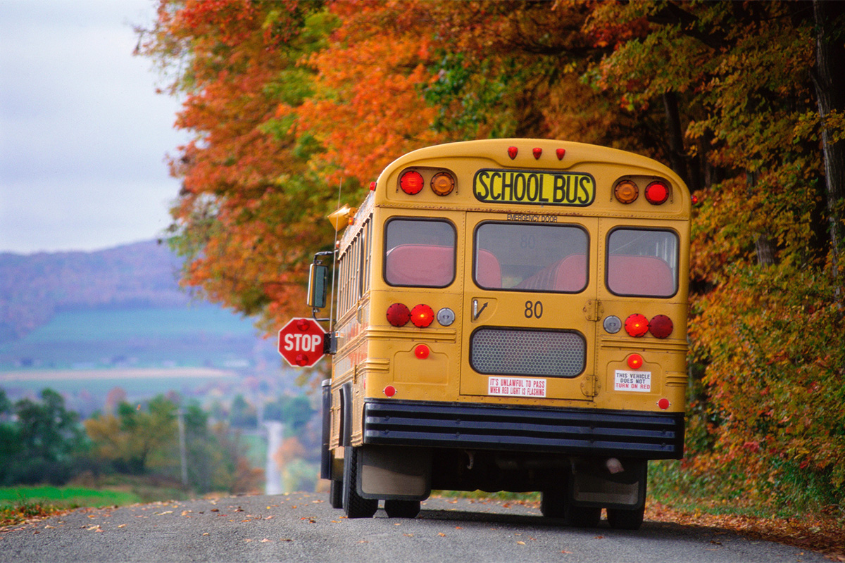 A yellow school bus stops, with its 'Stop' sign visible, along a country road in autumn."