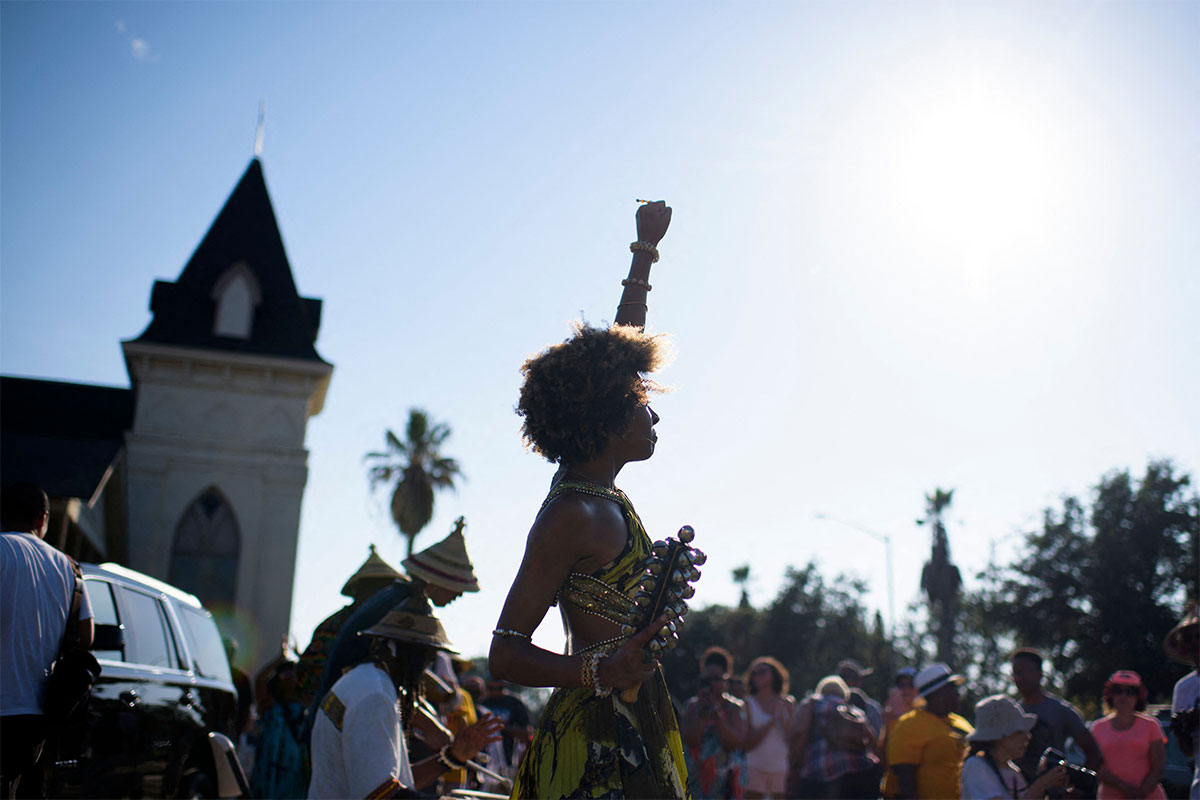 with a blue sky in the background, a Black woman stands over a crowd of people, raising her fist in the air.