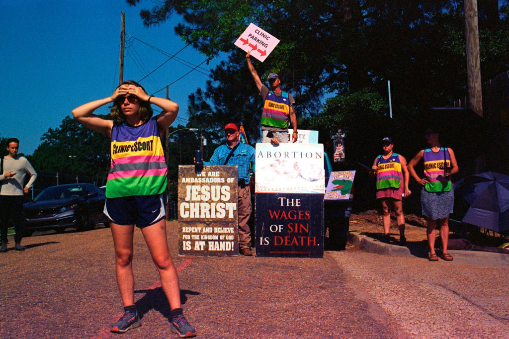 A clinic escort holds her hands over her forehead, looking in the distance, while anti-abortion protesters with grotesque signs depicting dead fetuses standing behind her, one saying "We Are Ambassadors For Jesus Christ." Behind the protesters, a man waves a sign that says "Clinic Entrance" with an arrow pointing right