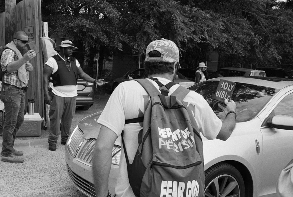 a photo shows a man with a backpack that says "Fear God" holding a Holy Bible up to a car, another man on the opposite side of the car is also holding a bible out to the car