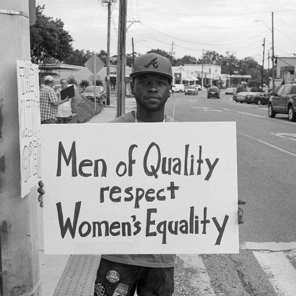 a photo shows a man holding a sign that says, "Men of Quality Respect Women's Equality"