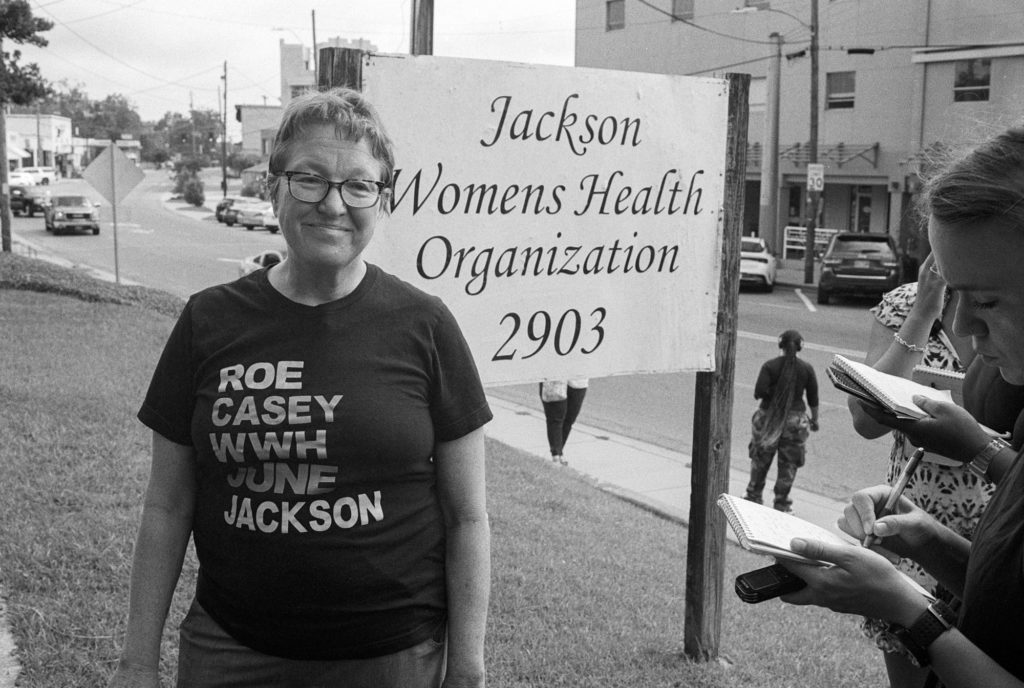 a woman stands outside the Jackson Women's Health Organization sign, wearing a black shirt that says "Roe, Casey, WWH, June, Jackson"