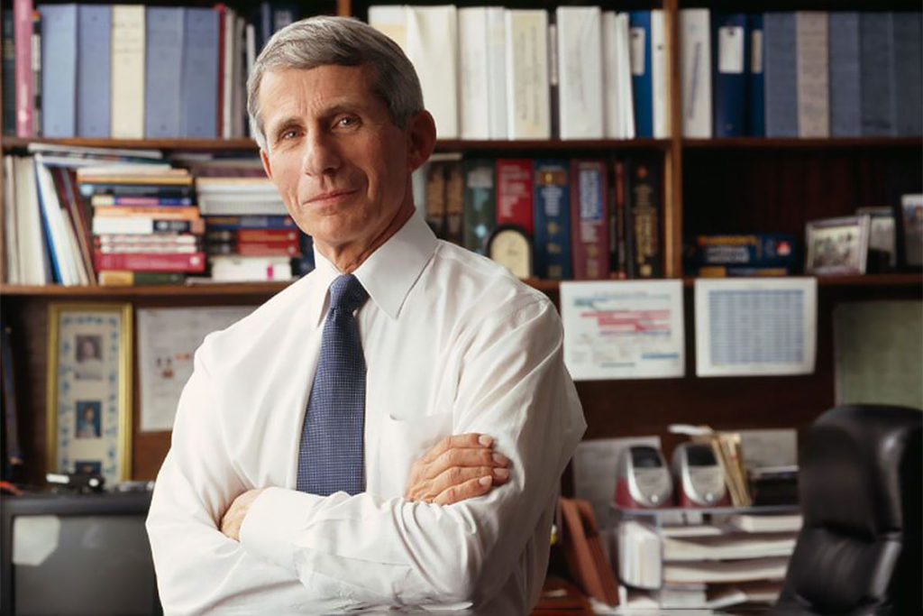 A grey haired man stands in a confident arms crossed pose in front of a cluttered office