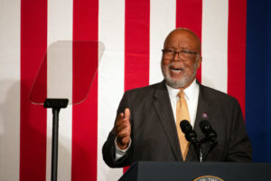 a photo shows Rep. Bennie thompson wearing a suit and tie and speaking into a microphone while looking at a glass teleprompter, the red and white stripes of an American flag hanging behind him