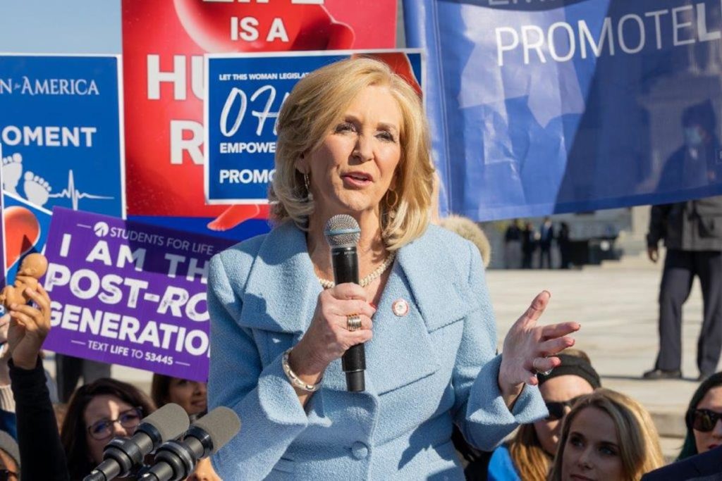 a photo of a woman holding a microphone with anti-abortion signs behind her, one says, "I Am the Post Roe Generation"