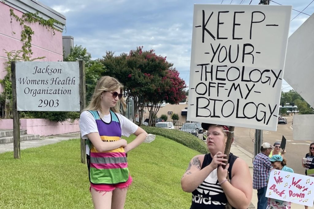 a photo of a clinic escort standing outside the pink building and the sign "Jackson Women's Health Organization" next to a protestor holding a sign that says, "Keep Your Theology Out Of My Biology"