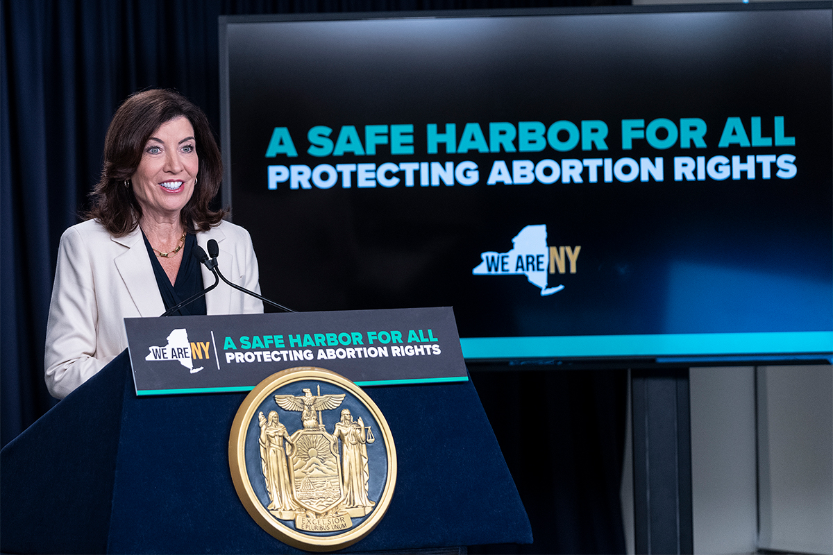 Kathy Hochul stands at a podium, in front of a screen that says 'A safe harbor for all: Protecting abortion rights.'