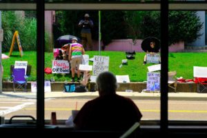 a photo shows the silhouette of a man sitting in a window with a drink and straw in front of him, gazing across the street at pro-choice protestors sitting on the grass in front of a pink building. Signs say, "My Body My Choice" and "Keep Your Theology Off My Body." The clinic wants the Mississippi Supreme Court to let it reopen