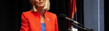 a photo of Lynn Fitch standing at a podium wearing a blue dress with a red coat over it