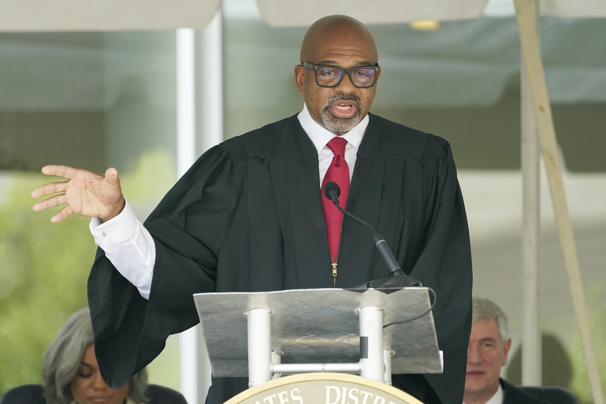 a photo of a Black man in a judge's dress wearing glasses and holding his hand up at a podium