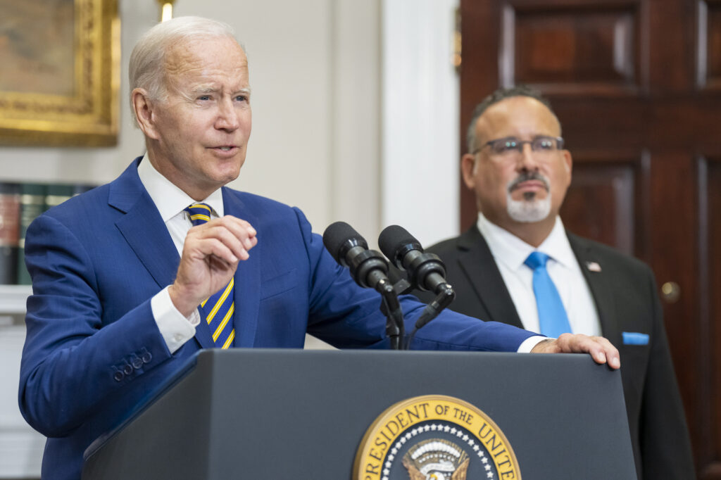 a photo of President Joe Biden standing at a podium in the White House with the education secretary standing behind him