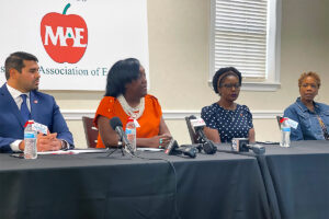 Four people sit at a long table with mics in front of them, and a sign for the Mississippi Association of Education behind them