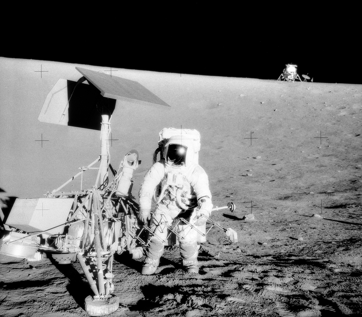 A person in a spacesuit standing next to a surveying craft on the surface of the moon with a lander in the background.