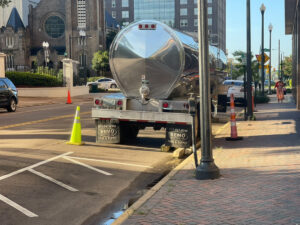 a photo shows a tanker truck parked on the side of a street next to a sidewalk across the street from a white brick wall with a black iron gate around it