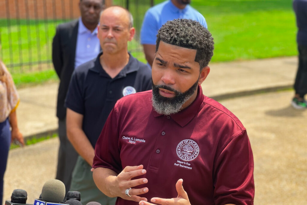 a photo of Chokwe Lumumba in a maroon shirt speaking in front of a gaggle of microphones with people standing behind him