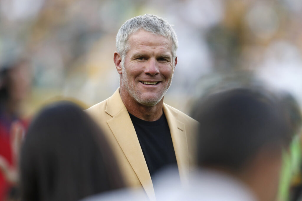 a photo of Brett Favre wearing a tan blazer over a black t-shirt in front of a crowded stadium