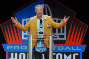 A man in a yellow NFL jacket spreads his arms wide at a clear podium at the Pro Football Hall of Fame
