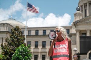photo shows a woman in a pink vest with the words "PINK HOUSE DEFENDERS" on it speaking into a megaphone while standing in front of the Mississippi Capitol building