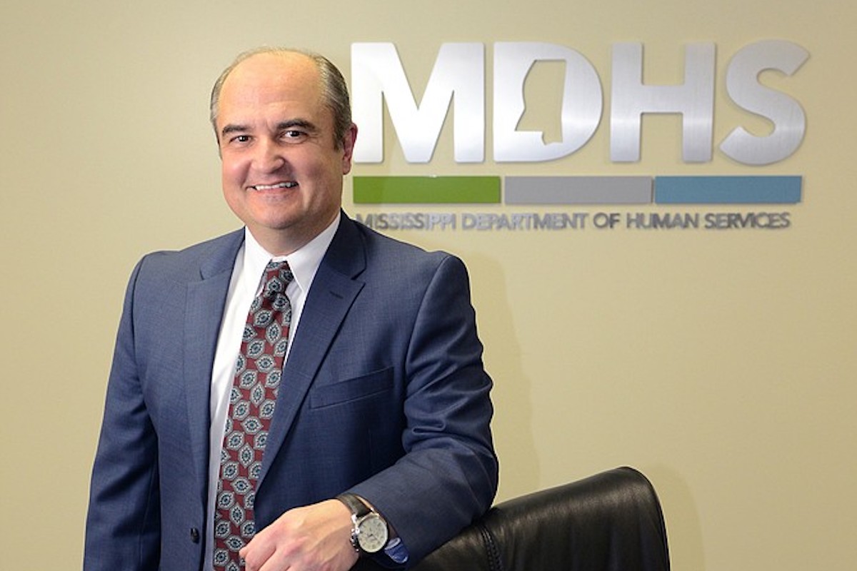 a photo of John Davis, a man with a balding head in a blue suit and tie in front of a sign that says MDHS