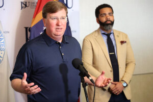 a photo of Tate Reeves speaking to a room with his arms out in a questioning way, and Chockwe Lumumba standing behind him, hands clasped