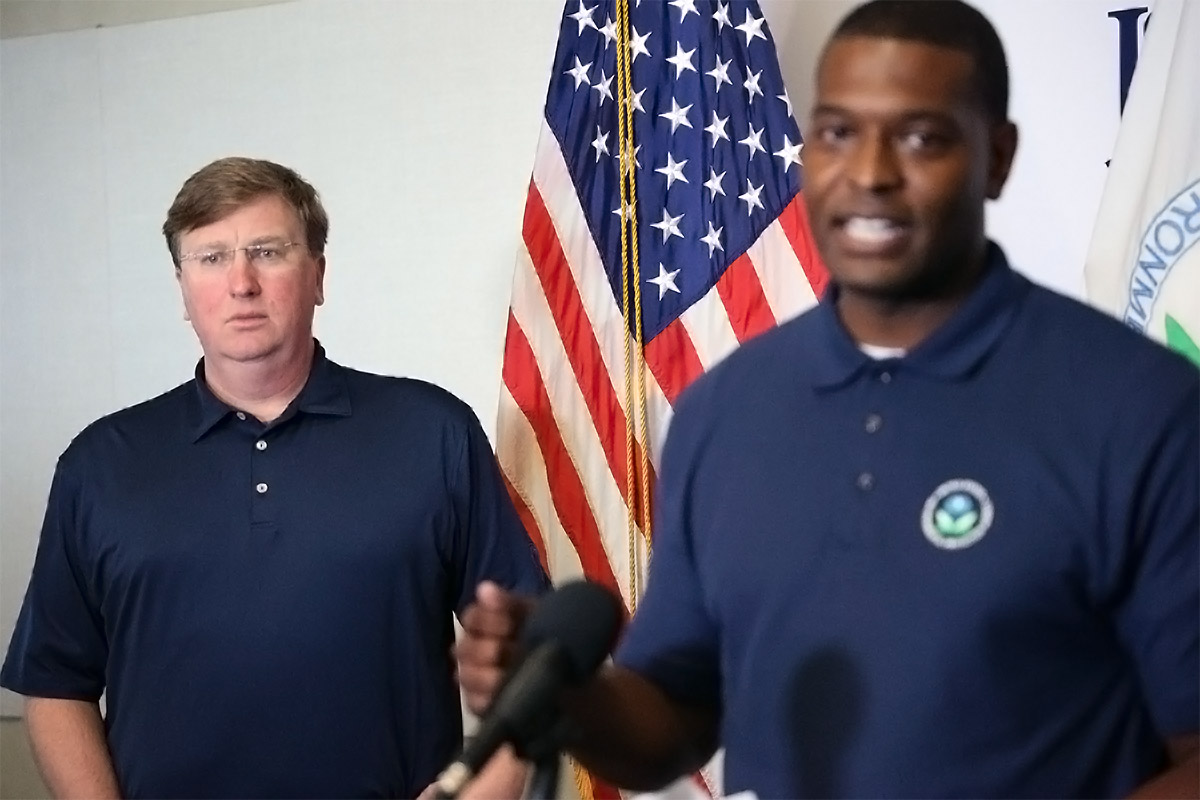 A man in the foreground speaks at a podium while a man in the back looks off in the distance