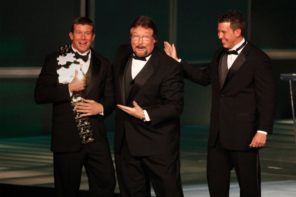 Three men in tuxedos on stage congratulating each other while the one on the left holds a trophy