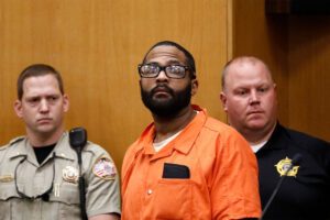 A man in a orange prison jumpsuit is flanked by two police officers in a courtroom