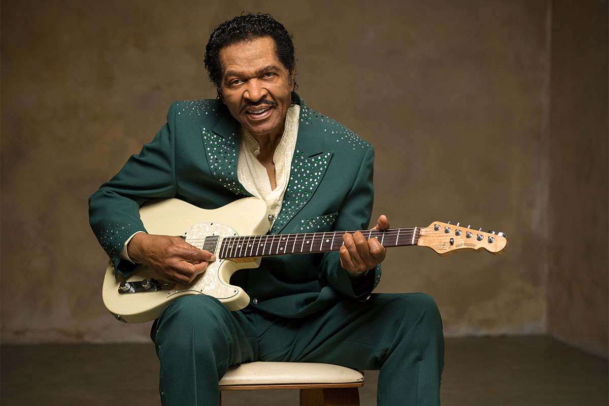Bobby Rush sitting down in a green suit playing a white bodied guitar