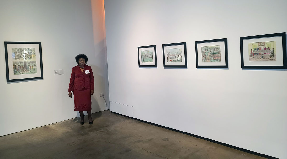 Gloria Gipson Suggs in a red suit dress standing in a gallery of her art