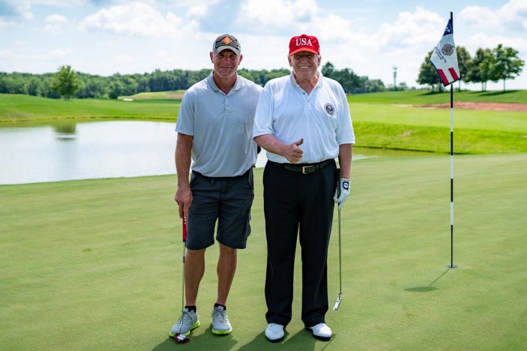 a photo of Brett Favre and Donald Trump standing side-by-side next to a golf hole in front of a pond, both holding golf clubs. Trump is wearing a red "USA" hat and holding a thumb up