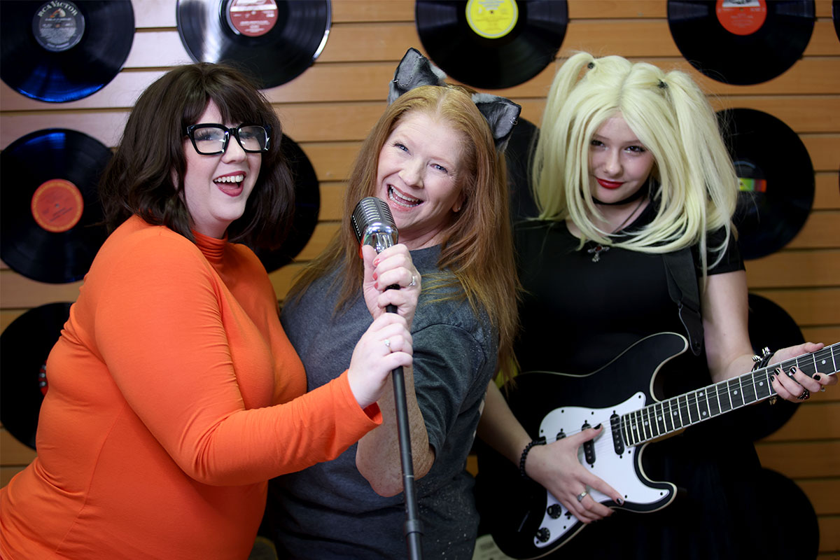 Three women dressed in costumes sing and play music in front of a wall filled with records