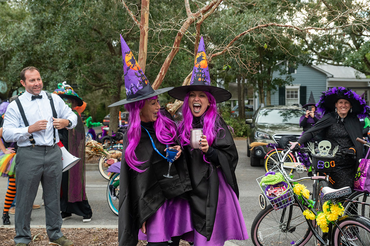 Two women dressed as witches in purple and black laugh in the center of the frame, with another witch with a bike off to the right