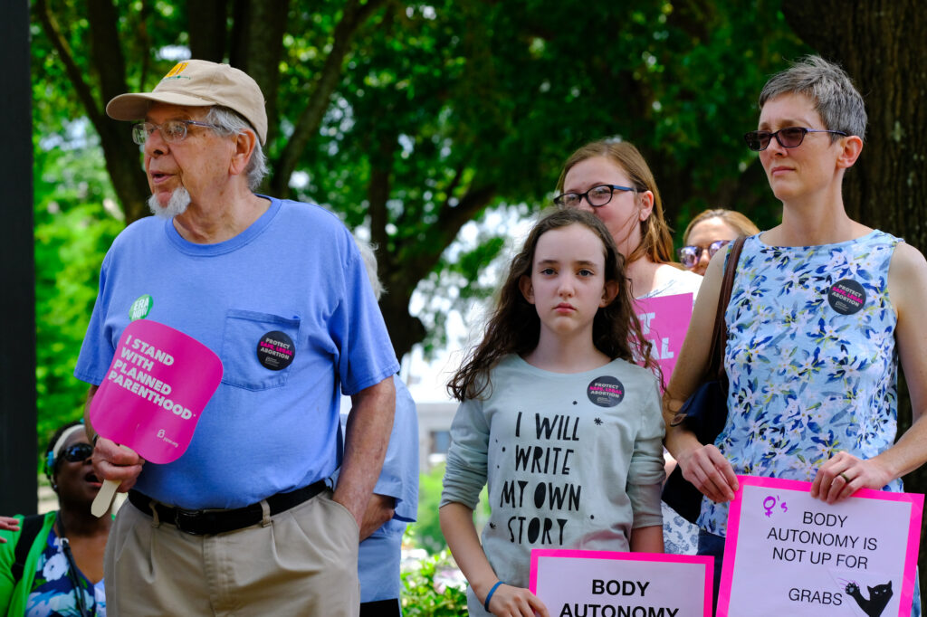 a photo shows three people holding signs supporting abortion rights, including a man, a woman, and a girl wearing a shirt that says "I will write my own story"
