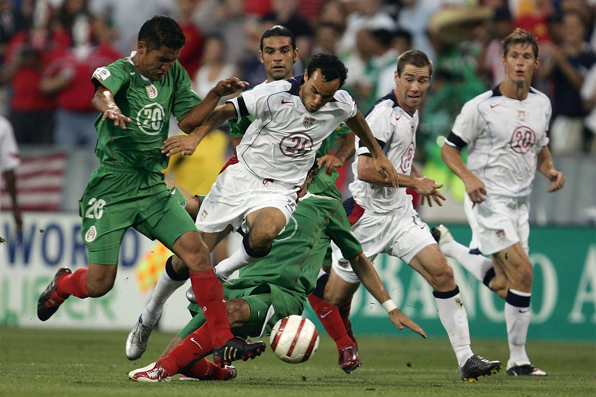 A footballer in a white jersey gets tackled by a player in a green jersey.