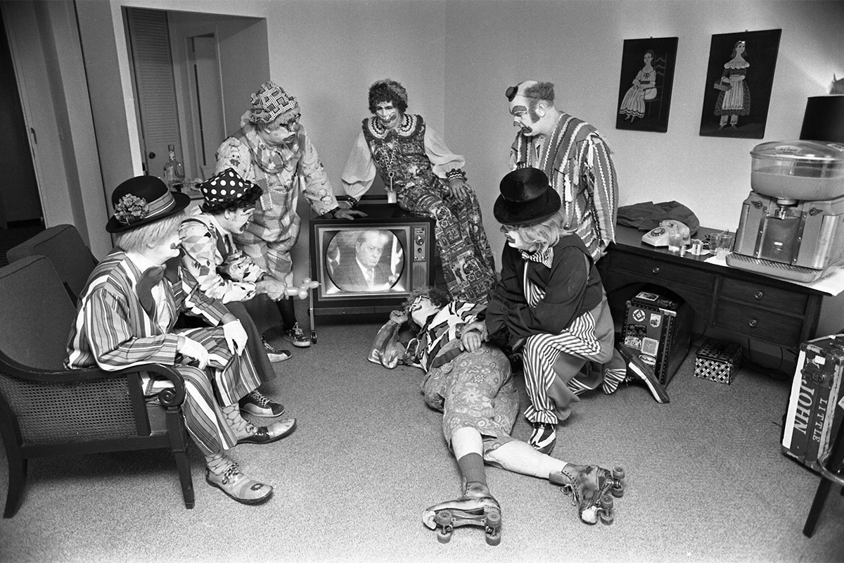 Many circus clowns in a room watching a TV.