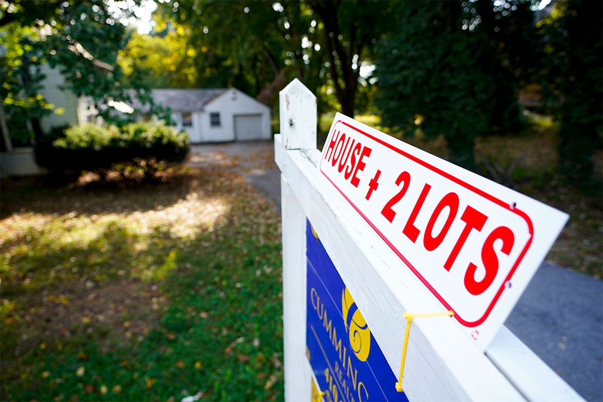 a for sale sign hangs on a white post in front of a home pictured in the distance