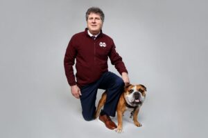 a photo of Mike Leach in a maroon MSU zip up kneeling next to a bulldog