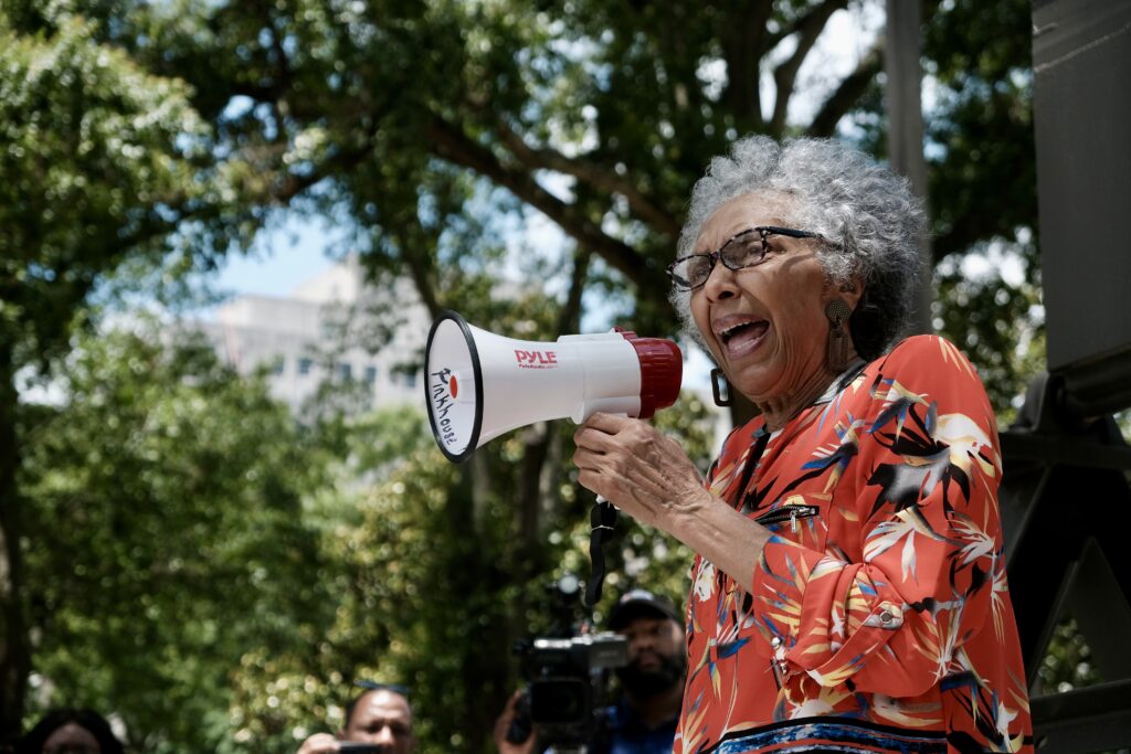 a photo shows a Black woman with gray hair and glasses standing outdoors holding a bullhorn and speaking