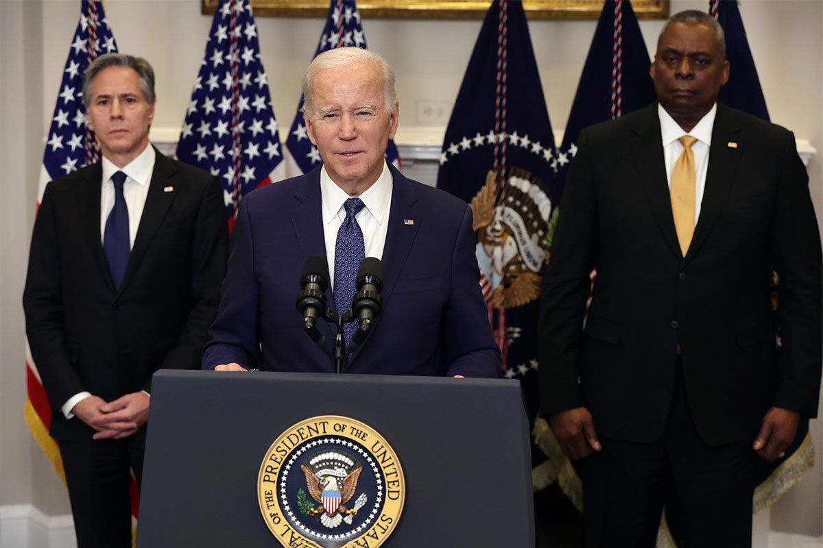 An older white man wearing a suit stands at a podium with the presidential seal on it . A middle aged black man and white man, both in suits, stand behind him, with American flags in the background