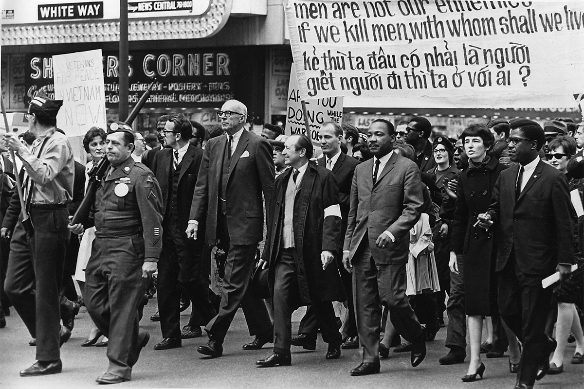 Led by Martin Luther King Jr., several men dressed in black suits march in a rally. Ahead of them are police officers holding rifles