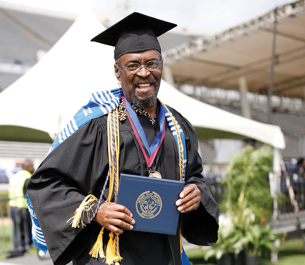 a photo shows a black man wearing a graduation cap and regalia while holding a diploma and smiling