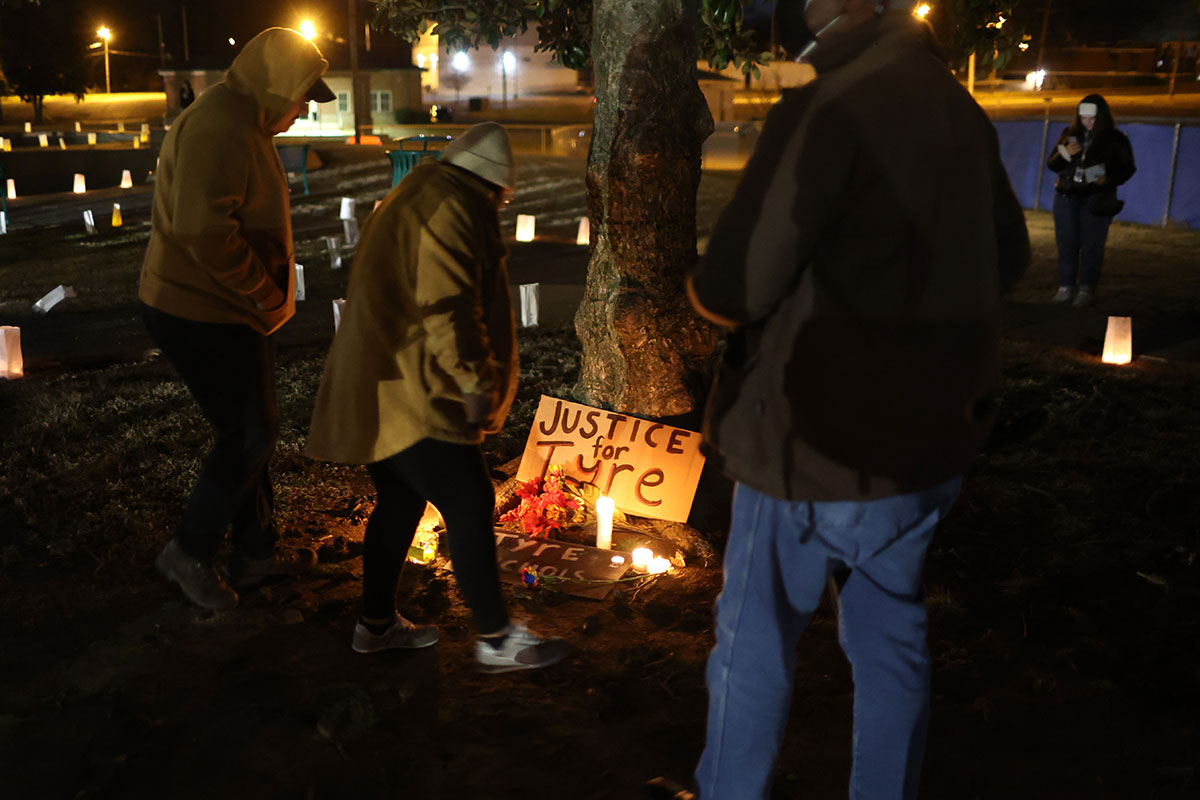 At night, three people wearing coats stand before a sign that is leaning against a tree and lit by candles. It reads, 'Justice for Tyre.