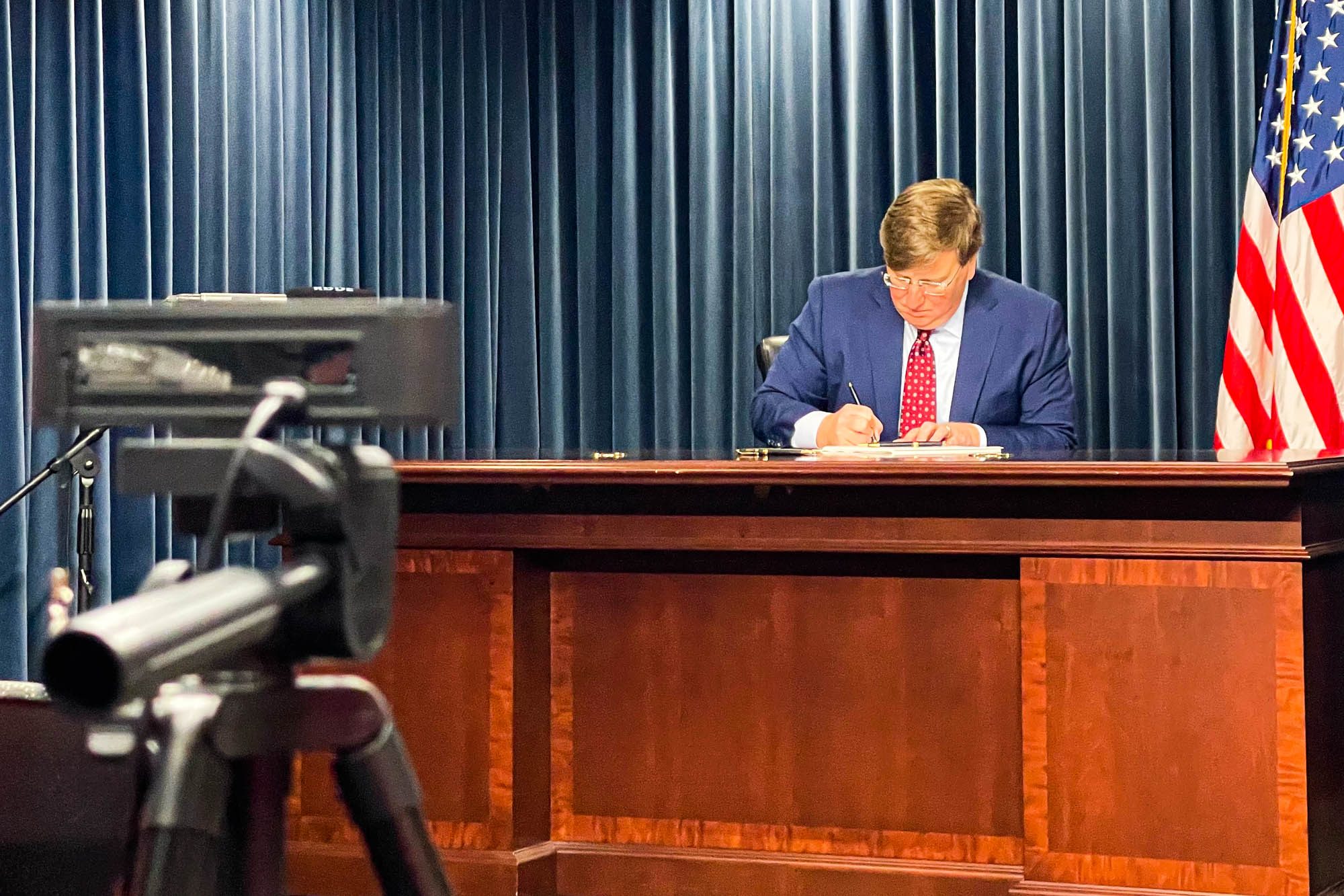 a photo of Tate Reeves at a desk signing a bill, with a camera in front of him