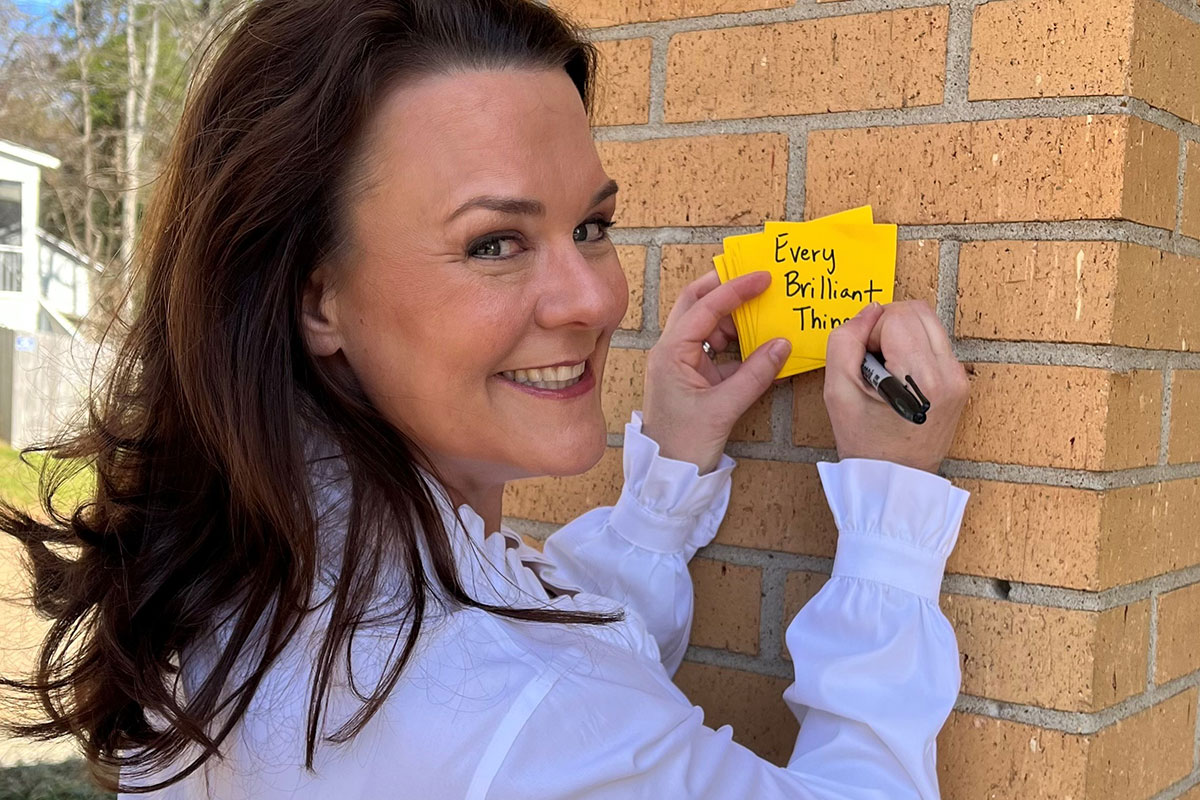 Ali Dinkins looks over her shoulder at the camera while writing "Every Brilliant Thing" on yellow post it notes