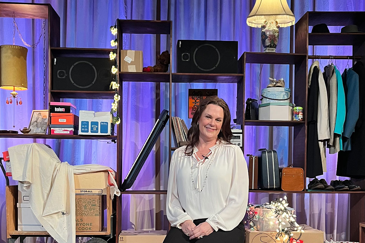 Ali Dinkins sits in a chair on stage while wearing a white top. The stage behind her has shelves full of household items