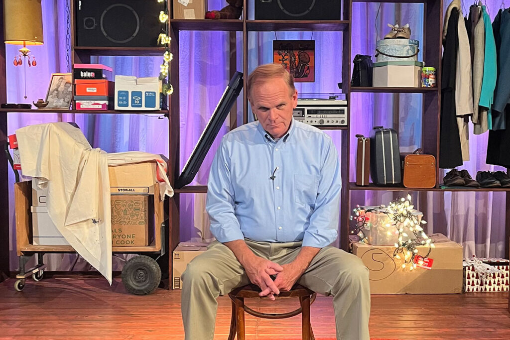 Ray McFarland in a blue button down and khaki pants sits in a chair on a stage