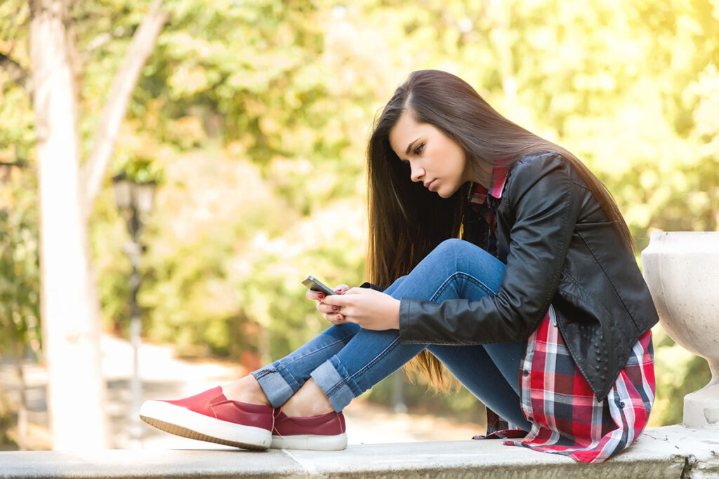A teenage girl in jeans and a black leather jacket sits outside while looking at a cell phone
